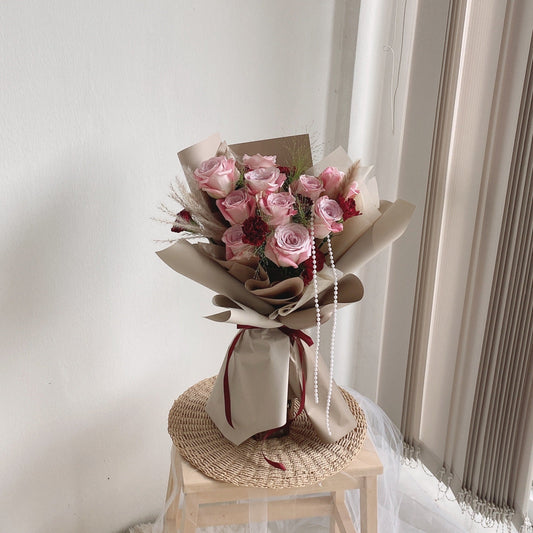 A bouquet of gooseberry pink roses wrapped in beige paper, displayed on a wooden platform with a note attached.