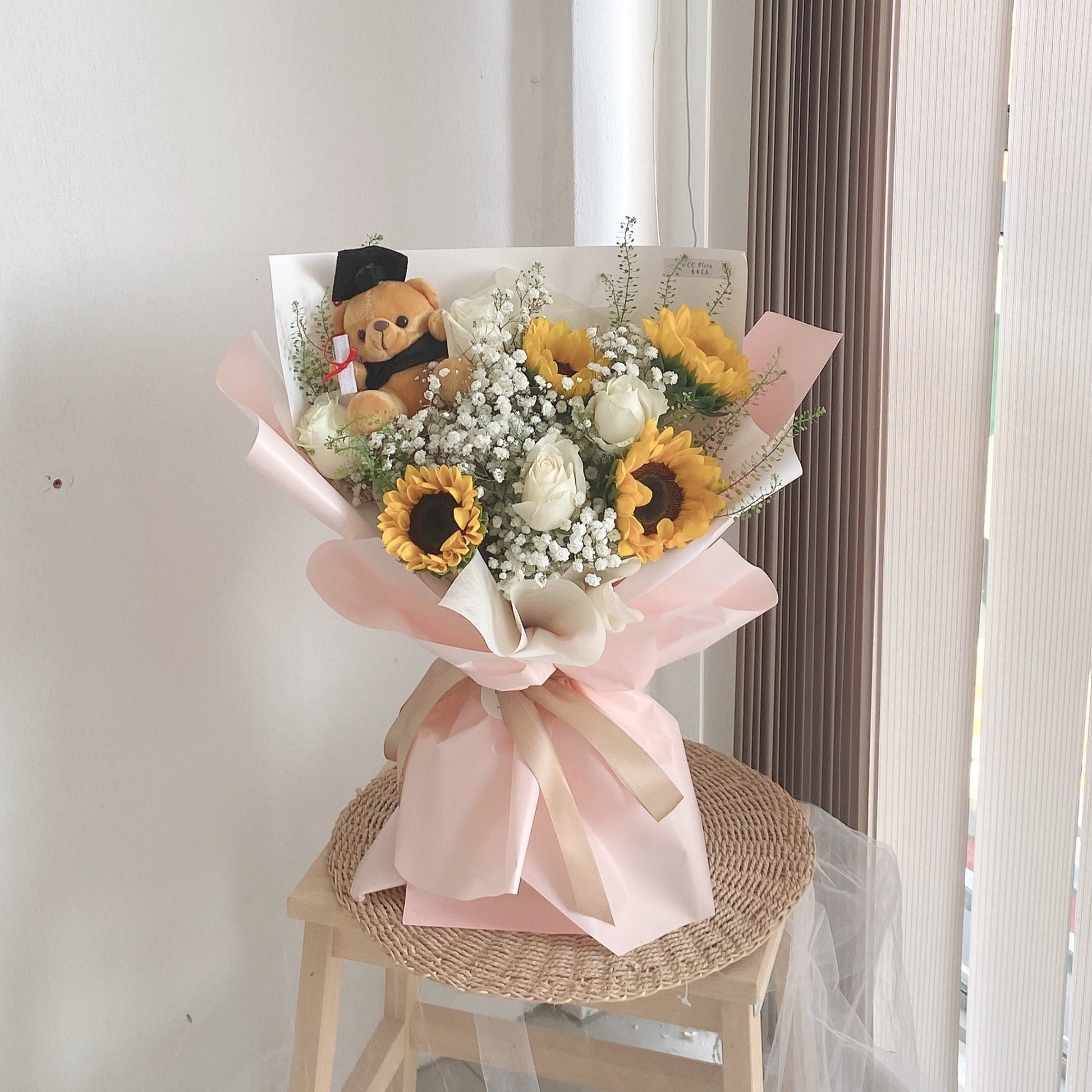 A bouquet of sunflowers, roses, and baby's breath with a teddy bear, wrapped in pink and white paper, displayed on a small table by a window.